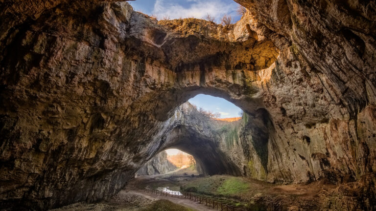 An awe-inspiring 4K wallpaper showcases the grand interior of Devetashka Cave in Devetaki, Bulgaria, defined by its spectacular natural arches and vast skylights. Golden sunlight streams dramatically through the immense ceiling openings, highlighting the textured rock walls and a winding path below.