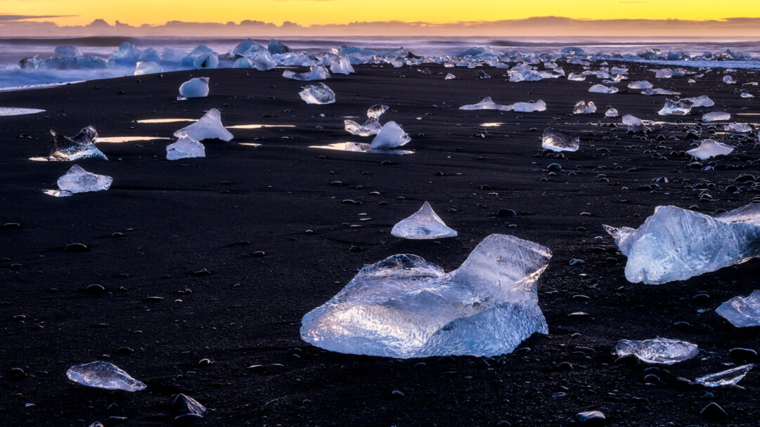 A captivating 4K wallpaper presents Diamond Beach in Iceland, where countless icebergs scatter across the black volcanic sand at sunset. The golden-orange sunset light bathes the distant ocean and horizon, while brilliant blue-white ice fragments sparkle against the dark, wet sand, creating a dramatic interplay of color and texture.