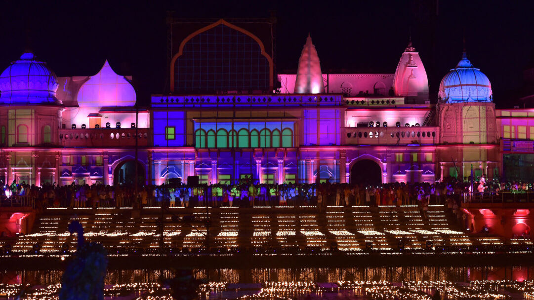 An enchanting 4K wallpaper showcases the festive Diwali celebrations along the Sarayu River banks, centered around a grandly illuminated temple. Thousands of flickering Diwali lamps adorn the river steps and float gracefully on the dark water, their warm glow beautifully reflecting the temple's vibrant purple, pink, and blue lighting.