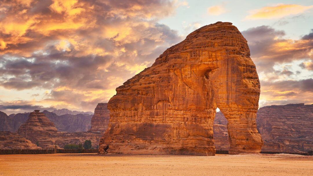 An epic 4K wallpaper showcases Elephant Rock in Al-Ula, Saudi Arabia, standing majestically in a vast desert landscape. The warm sunset light bathes the towering, elephant-shaped sandstone formation and dramatic, fiery orange clouds, creating a breathtaking and serene desert mood.