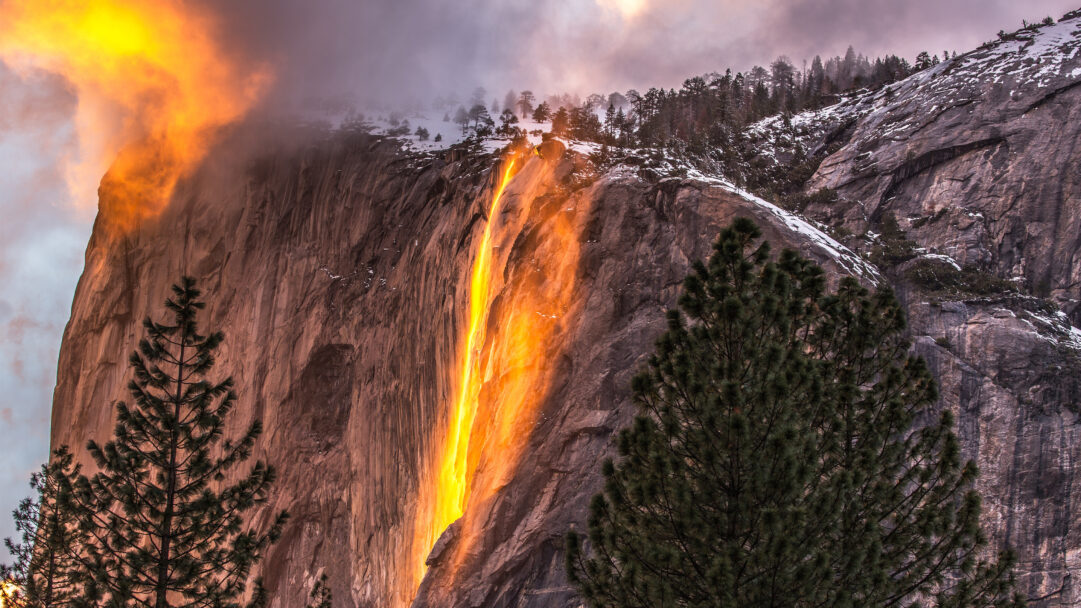 A breathtaking 4K wallpaper capturing the famous Firefall event at Horsetail Fall in Yosemite National Park, California, where the waterfall plunges down a massive granite formation. The ephemeral phenomenon illuminates the cascading water in brilliant fiery orange and yellow hues, creating a stunning visual of liquid light against the shadowed, snow-dusted cliffs and misty sky.