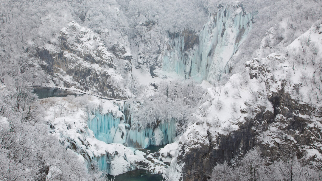 A breathtaking 4K wallpaper showcasing the cascading frozen waterfalls within the snowy canyon of Plitvice Lakes National Park, Croatia, during winter. Vibrant turquoise ice formations cling dramatically to the snow-covered cliffs, with touches of dark water contrasting the pristine winter landscape.