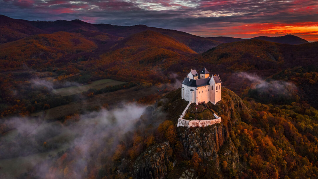 A dramatic 4K wallpaper showcasing Füzér Castle perched majestically in the Zemplén Mountains, Hungary, during autumn. The castle is illuminated against the vibrant orange and crimson sunset sky, with swirling fog partially obscuring the surrounding autumn-hued forests, creating a mystical atmosphere.