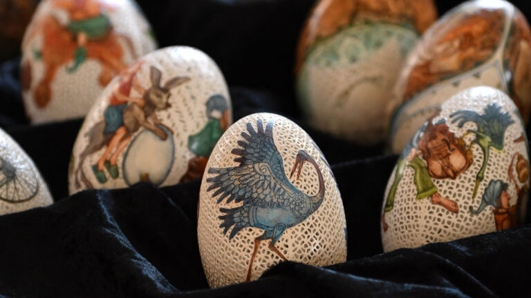 An exquisite 4K wallpaper showcasing a collection of hand-painted carved Easter eggs by Tünde Csuhaj from Szekszárd, Hungary, delicately displayed on a dark, plush fabric. The delicate, lace-like carving on each egg beautifully frames the vibrant, hand-painted scenes, creating a captivating interplay of light and texture that highlights the intricate artistry.