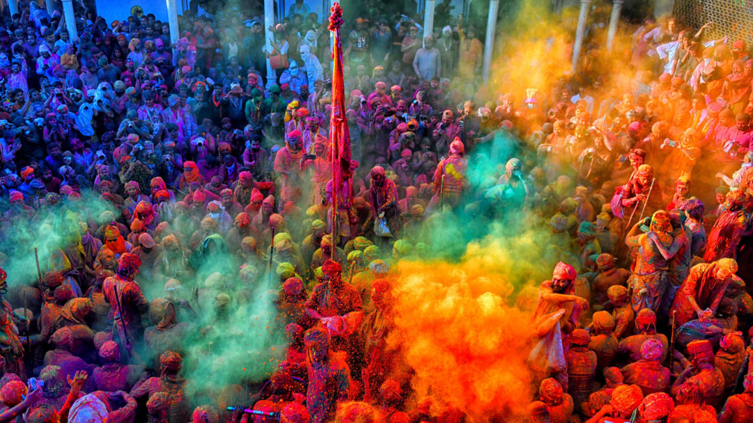 An electrifying 4K wallpaper capturing a dense crowd joyfully celebrating Holi in Mathura, Uttar Pradesh, India. Vivid plumes of red, orange, green, and blue powder explode through the air, completely engulfing the jubilant participants in a breathtaking spectacle of color and festive energy.