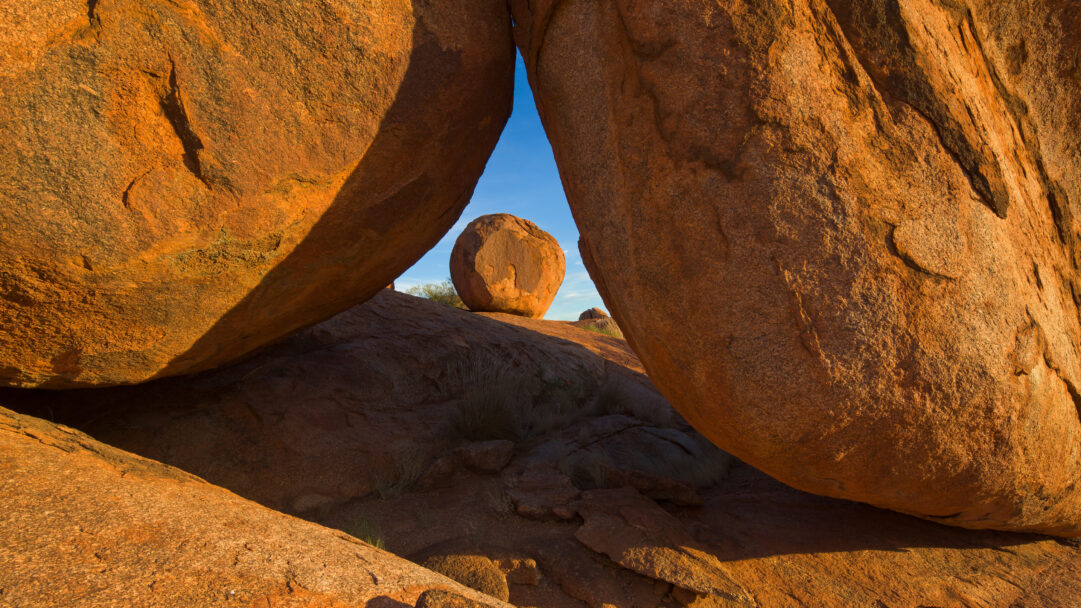 A captivating 4K wallpaper showcasing the iconic Karlu Karlu Devils Marbles in Australia, where massive orange boulders dominate a desert landscape. Golden sunlight dramatically illuminates their weathered surfaces, highlighting the striking spherical boulder perfectly framed through an arching crevice against a vibrant blue sky.