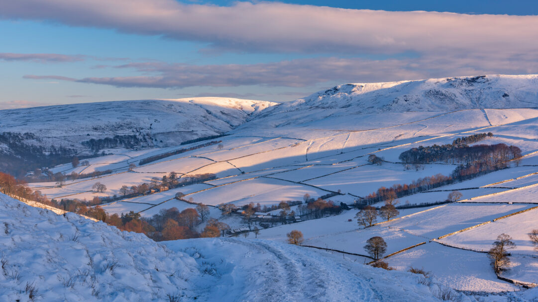 A magnificent 4K wallpaper unveils the Kinder Scout winter landscape across the snow-draped Peak District at sunrise. Golden light dramatically illuminates the undulating snow-covered slopes, casting deep blue shadows that delineate fields and valleys, evoking a serene yet crisp winter morning.
