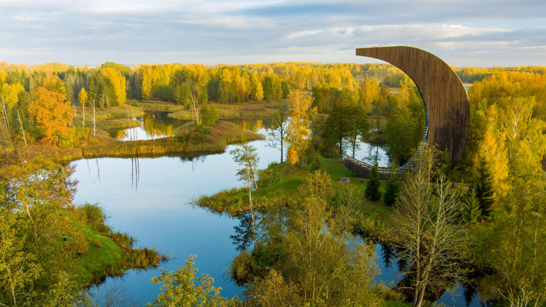 A breathtaking 4K wallpaper captures the Kirkilai Lakes Lookout Tower overlooking an expansive autumn landscape within Biržai Regional Park, Lithuania. The unique crescent-shaped tower stands amidst numerous calm lakes and forests ablaze with brilliant golden and orange foliage under a soft, diffused sky.