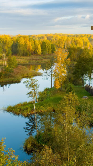 A breathtaking 4K wallpaper captures the Kirkilai Lakes Lookout Tower overlooking an expansive autumn landscape within Biržai Regional Park, Lithuania. The unique crescent-shaped tower stands amidst numerous calm lakes and forests ablaze with brilliant golden and orange foliage under a soft, diffused sky.