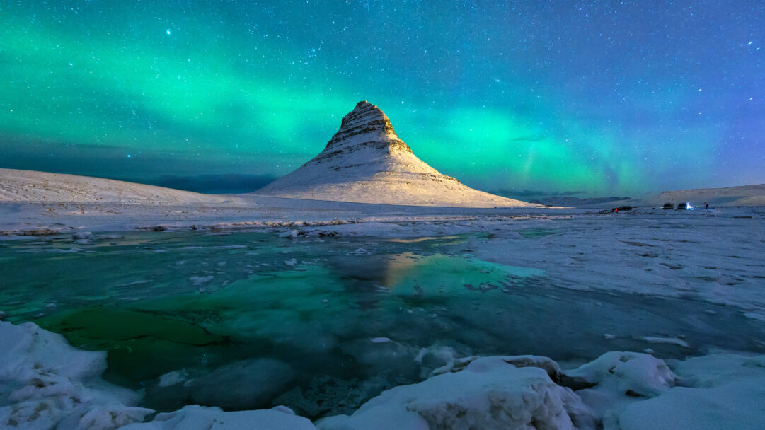 An ethereal 4K wallpaper of Kirkjufell Mountain, snow-covered and majestic, standing in a serene winter Icelandic landscape under the swirling Aurora Borealis. The mountain's illuminated peak and the vivid green-blue Northern Lights are mirrored flawlessly in the still, partially frozen water, enhancing the scene's breathtaking magic.