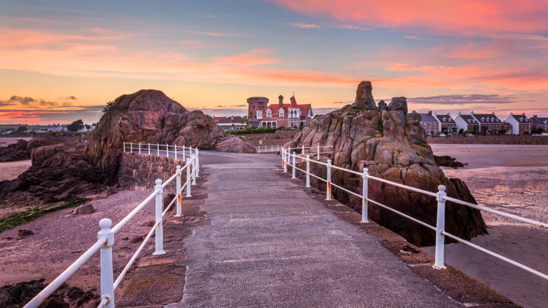 An enchanting 4K wallpaper depicts La Rocque Harbour in St. Clement, Jersey, at sunset, where a coastal path winds between dramatic rock formations. The sky blazes with soft pink and orange hues, casting a warm glow over the textured coastal rocks and the expansive sandy beach, creating a serene and inviting atmosphere.