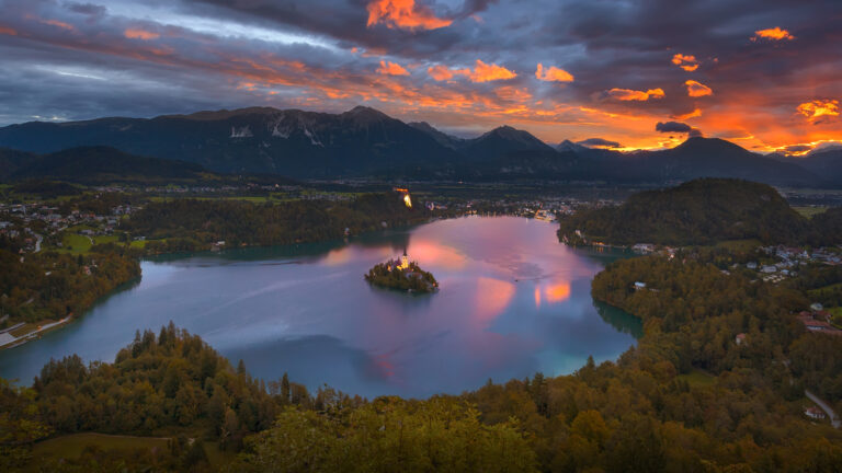 A breathtaking 4K wallpaper showcasing Lake Bled in Slovenia at sunrise, with its iconic island church and Bled Castle prominently featured against a mountainous backdrop. The sky is ablaze with vibrant orange and purple clouds, their dramatic colors reflected perfectly in the tranquil lake, creating a serene and majestic mood.