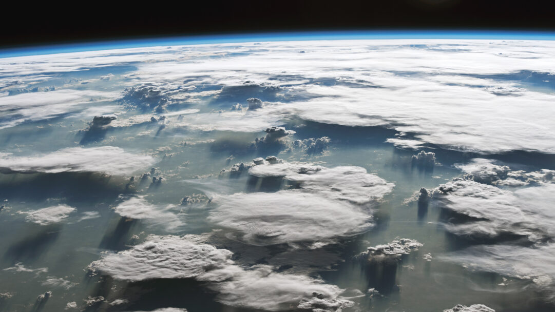 A grand 4K wallpaper displays immense anvil clouds spanning over the Amazon basin in Brazil, as seen from a high Earth orbit. Dramatic sunlight streams through breaks in the clouds, creating long, dark shadows that stretch across the vast, lighter cloud cover and atmospheric haze below, emphasizing the depth and scale of the weather system.