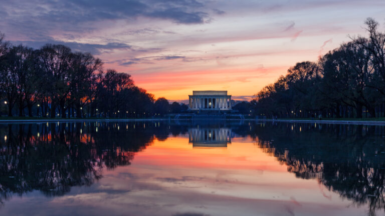 An evocative 4K wallpaper presenting the Lincoln Memorial grandly situated at the end of the Reflecting Pool in Washington DC, bathed in the soft glow of a sunset. The vivid orange, pink, and purple hues of the setting sun are spectacularly mirrored in the still water, creating a symmetrical and tranquil scene.