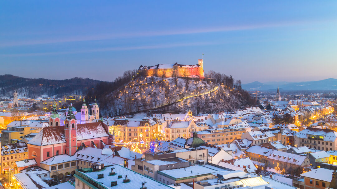 A breathtaking 4K wallpaper presenting Ljubljana Castle majestically illuminated atop a snow-covered hill in a winter cityscape of Slovenia at night. The castle and festive street lights cast a warm glow across the pristine snow-dusted rooftops, creating an enchanting and serene twilight atmosphere.