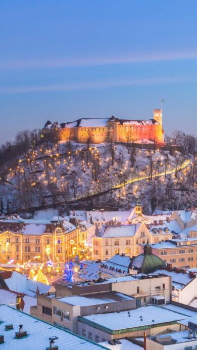 A breathtaking 4K wallpaper presenting Ljubljana Castle majestically illuminated atop a snow-covered hill in a winter cityscape of Slovenia at night. The castle and festive street lights cast a warm glow across the pristine snow-dusted rooftops, creating an enchanting and serene twilight atmosphere.