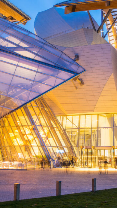 A captivating 4K wallpaper showcasing the Louis Vuitton Foundation in Paris at night. Its iconic, illuminated architecture features grand glass “sails” glowing with warm golden light, creating a striking contrast against the deep twilight sky and the blurred movement of visitors.
