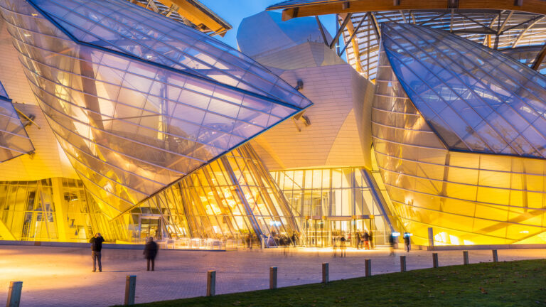 A captivating 4K wallpaper showcasing the Louis Vuitton Foundation in Paris at night. Its iconic, illuminated architecture features grand glass “sails” glowing with warm golden light, creating a striking contrast against the deep twilight sky and the blurred movement of visitors.