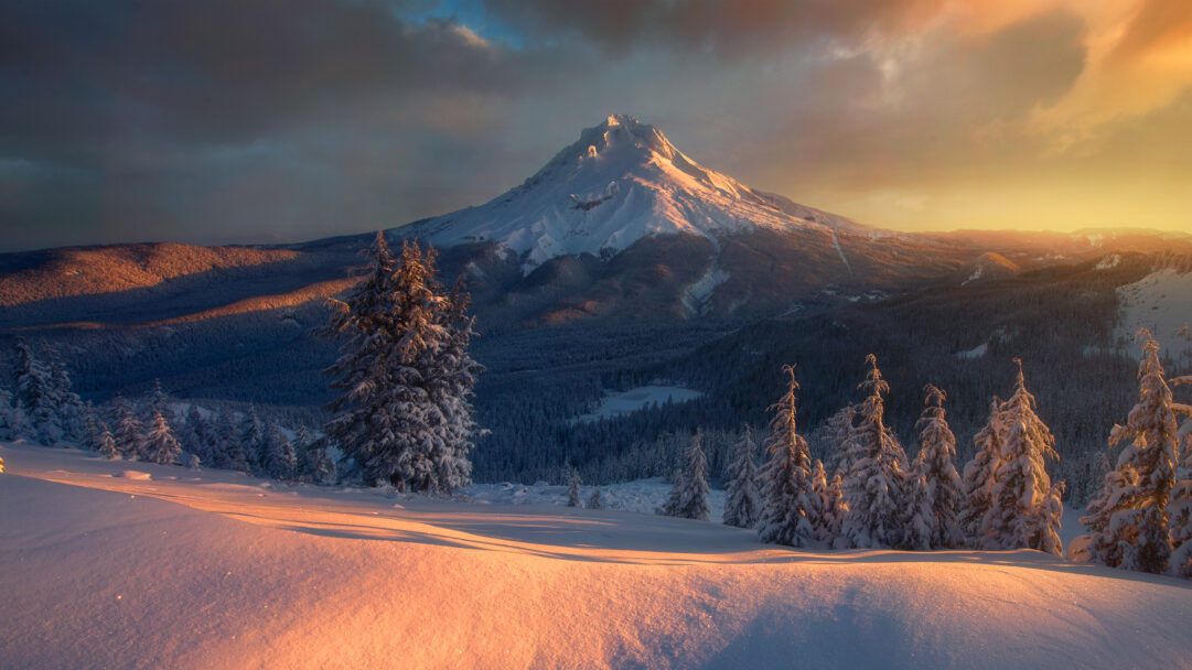 An awe-inspiring 4K wallpaper captures the majestic snow-capped Mount Hood presiding over a serene winter landscape in Oregon. Golden sunset light bathes the snow-dusted foreground and the mountain's peak, creating a dramatic interplay of warm oranges and cool blue shadows across the tranquil scene.