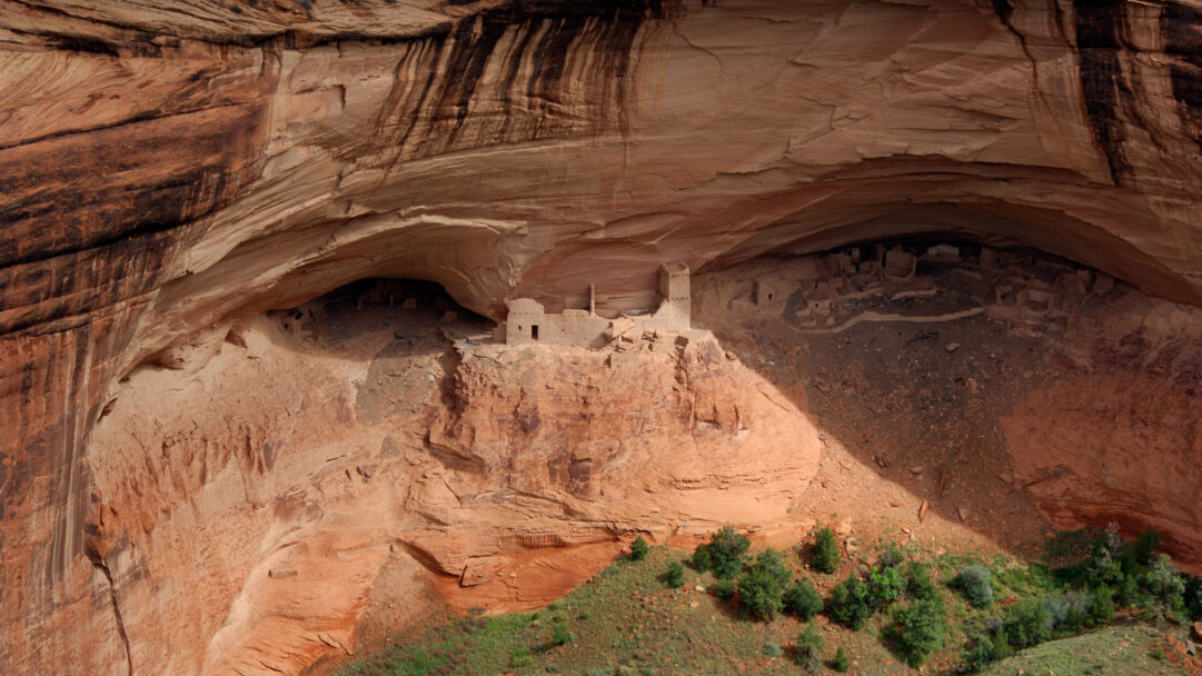 A majestic 4K wallpaper capturing the ancient Mummy Cave Ruins, an impressive collection of Ancestral Puebloan cliff dwellings nestled deep within the dramatic ochre cliffs of Canyon de Chelly National Monument, Arizona. Sunlight dramatically illuminates the central tower and a portion of the cliff face, highlighting the intricate textures of the sand-colored ruins and the stratified red rock, while deep shadows conceal other ancient structures, evoking a sense of enduring history and grandeur.