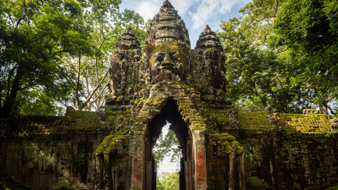 A majestic 4K wallpaper capturing the ancient North Gate of Angkor Thom Archaeological Park, a towering stone structure with multiple serene faces carved into its spires and an arched entryway, framed by lush tropical trees. Heavy green moss and creeping vines richly cover the weathered stone, emphasizing the gate's intricate details and imparting a profound sense of historical grandeur and jungle reclaim.