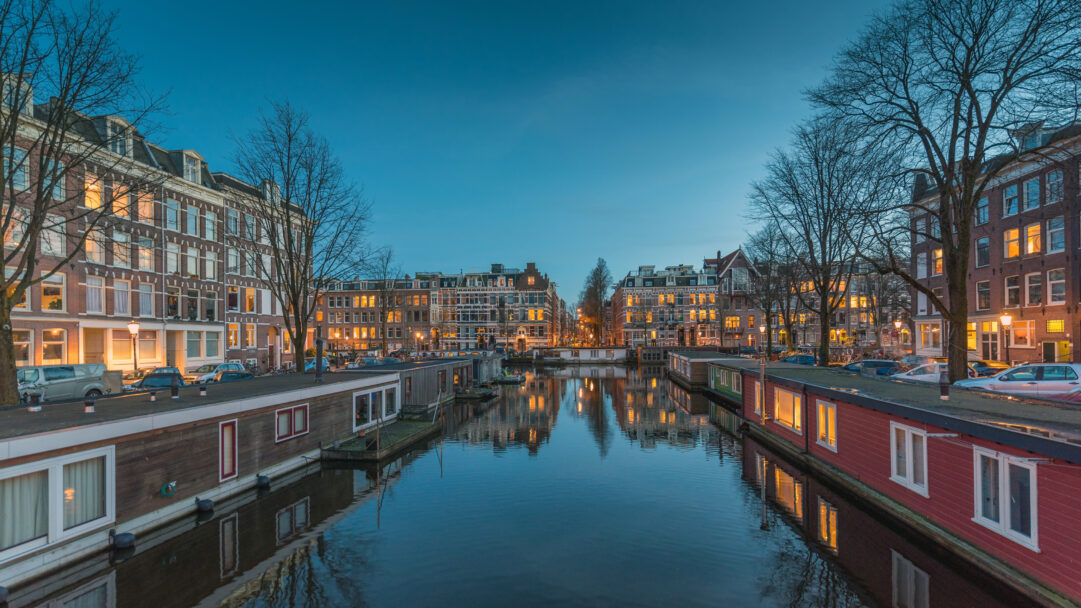 An enchanting 4K wallpaper capturing the Oud-West Amsterdam Canal at night, lined with houseboats and historic city buildings. Warm lights from the numerous windows and streetlamps create captivating reflections on the tranquil canal water, defining the peaceful evening mood.