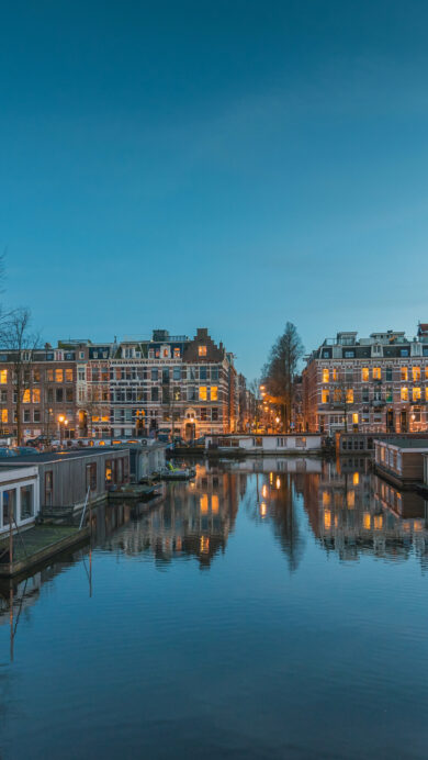 An enchanting 4K wallpaper capturing the Oud-West Amsterdam Canal at night, lined with houseboats and historic city buildings. Warm lights from the numerous windows and streetlamps create captivating reflections on the tranquil canal water, defining the peaceful evening mood.