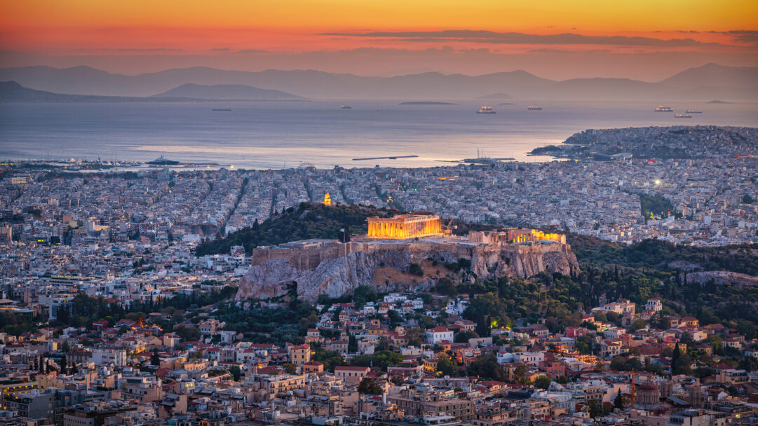 A magnificent 4K wallpaper showcasing a panoramic view of the Athens cityscape and the grand Acropolis at sunset in Greece, extending towards the tranquil Aegean Sea and distant mountains. The ancient structures of the Acropolis are beautifully illuminated in warm golden light, contrasting with the vibrant orange and deep blue hues of the twilight sky and the twinkling lights of the vast city below.