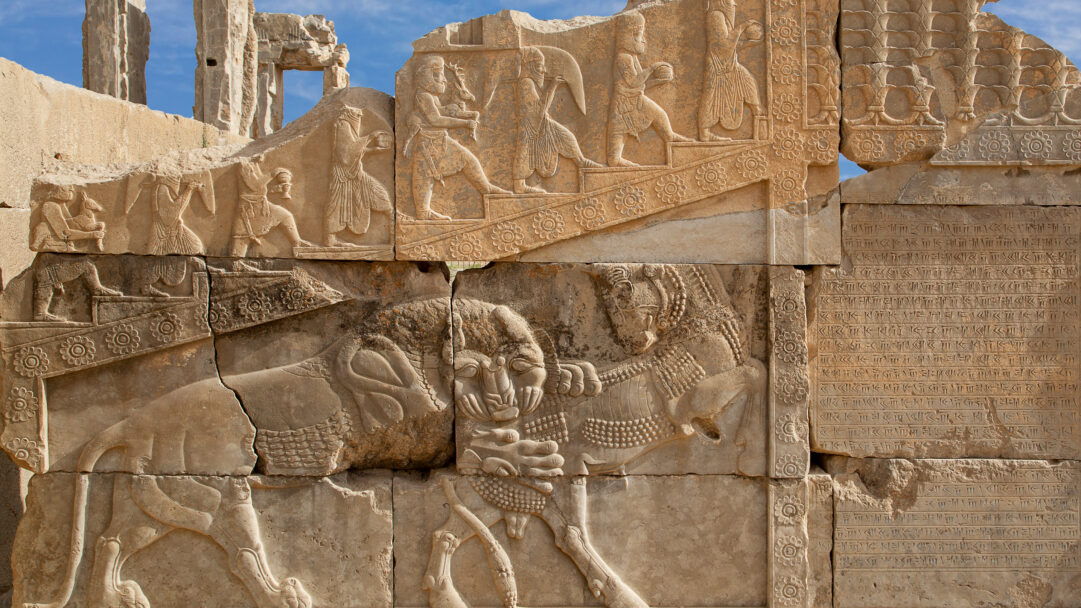 A majestic 4K wallpaper revealing ancient Persian stone reliefs at Persepolis, Iran, under a bright blue sky. Intricate carvings depict mythological struggles like a lion attacking a bull and figures ascending stairs with offerings, alongside ancient cuneiform script, all bathed in warm light that accentuates their weathered texture and historical depth.