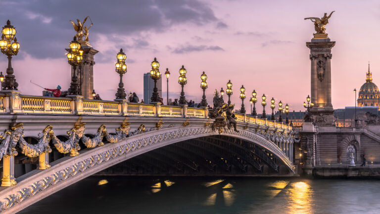 A majestic 4K wallpaper of the ornate Pont Alexandre III Bridge spanning the Seine River in Paris, France, at twilight. Its intricate golden statues and countless glowing lamps cast a warm, inviting glow against the soft pink and lavender twilight sky, reflecting enchantingly on the calm river below.