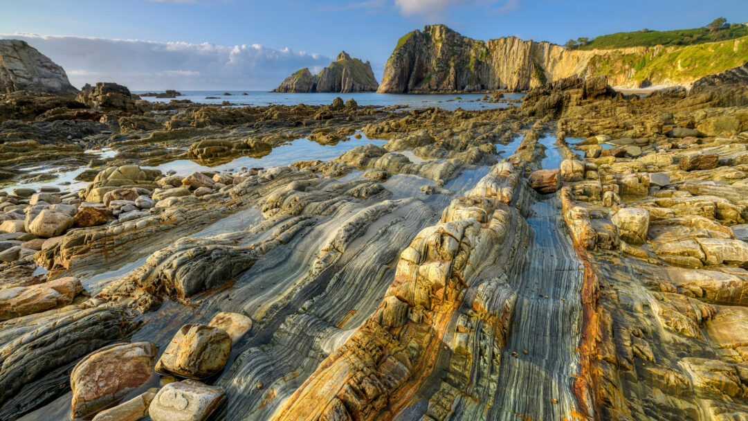 An awe-inspiring 4K wallpaper capturing the intricate quartzite formations dominating the foreground of Playa del Silencio on the Asturias, Spain coast. Golden hour light dramatically illuminates the deeply layered quartzite patterns in the foreground tidal pools, leading the eye towards the majestic sea stacks and towering cliffs under a clear blue sky.