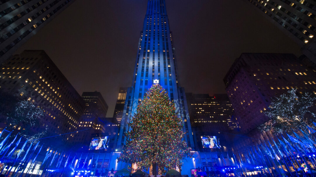 A magnificent 4K wallpaper showcasing the iconic Rockefeller Center Christmas Tree at night in New York City, towering amidst surrounding illuminated skyscrapers. Its myriad of colorful lights sparkle brilliantly against the deep blue facade of the main building and the dark winter sky, creating a truly festive and majestic holiday spectacle.