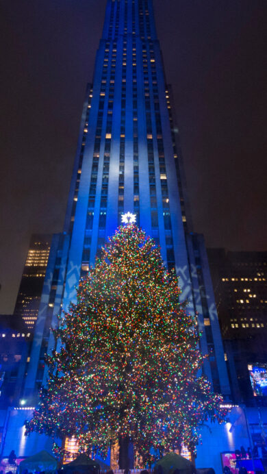 A magnificent 4K wallpaper showcasing the iconic Rockefeller Center Christmas Tree at night in New York City, towering amidst surrounding illuminated skyscrapers. Its myriad of colorful lights sparkle brilliantly against the deep blue facade of the main building and the dark winter sky, creating a truly festive and majestic holiday spectacle.