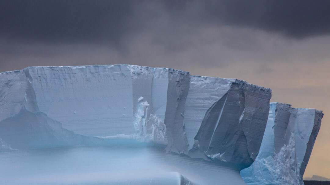 A colossal 4K wallpaper presents a massive iceberg towering dramatically in the vast, frigid landscape of Antarctica's Ross Sea. Its sheer, layered blue-white ice cliffs glow faintly against a moody sky, emphasizing the icy grandeur and desolate beauty of this polar environment.