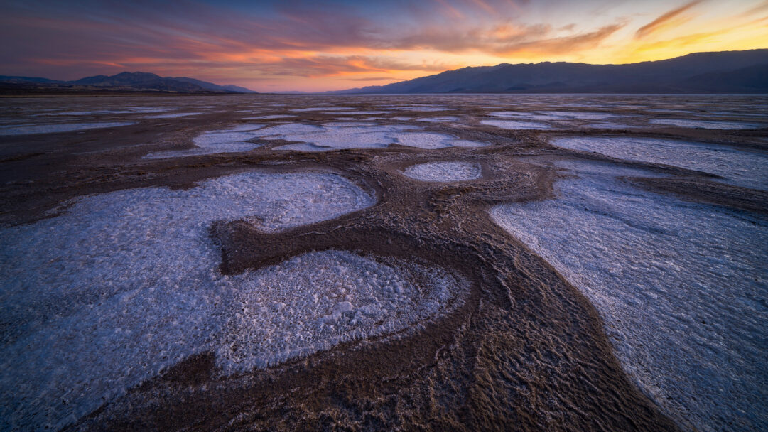 A mesmerizing 4K wallpaper of the expansive salt flats in Badwater Basin, Death Valley National Park, California. The striking geometric patterns of the white salt crust contrast dramatically with the dark basin floor under a radiant sunset sky of fiery orange, soft pink, and deep blue hues.