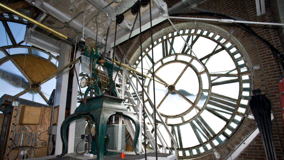 An intricate 4K wallpaper revealing the complex mechanical clock interior of the San Jacinto Building in Beaumont, Texas. Bright light streams through the large, gridded clock face, illuminating the gleaming brass gears and heavy chains that power this historical timepiece.