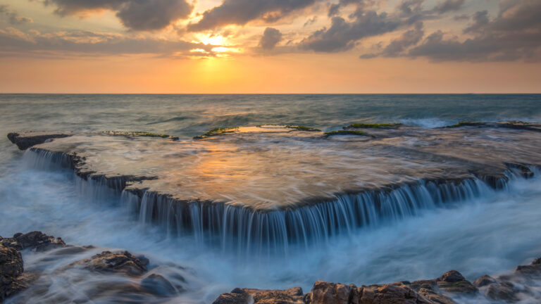 A captivating 4K wallpaper of seawater cascading over an ancient coral reef in Hang Rai, Vietnam, at sunset. The ethereal, long-exposure effect blurs the blue-green seawater into silky cascades, reflecting the fiery orange and yellow sunset across the reef's surface.