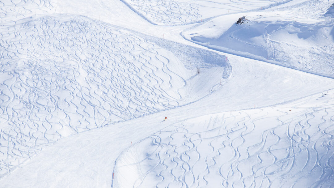 A breathtaking 4K wallpaper featuring a lone skier carving down the expansive, snow-covered slopes of Bernina Pass, Graubünden, Switzerland. The intricate patterns of fresh ski tracks blanket the pristine white landscape, with the vibrant orange jacket of the skier providing a solitary, energetic focal point.