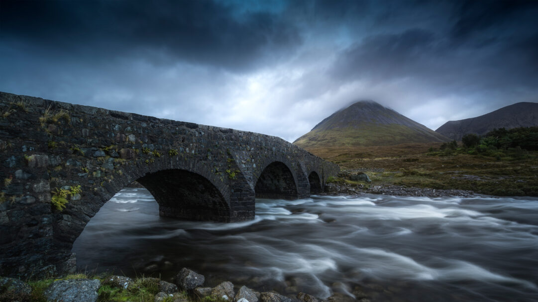 A dramatic 4K wallpaper of the Sligachan Old Bridge on the Isle of Skye, Scotland, majestically spanning a fast-flowing river with a prominent, mist-shrouded mountain rising in the background. The long-exposure effect blurs the river into a silky, white current, contrasting with the ancient stone bridge and the dark, atmospheric clouds overhead, evoking a sense of timeless Scottish wilderness.