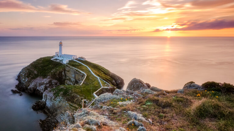 An awe-inspiring 4K wallpaper capturing the iconic South Stack Lighthouse perched on a rugged island cliff off Holy Island, Wales. The tranquil sea reflects the vibrant orange and pink hues of the sunset, creating a breathtaking and serene coastal landscape.