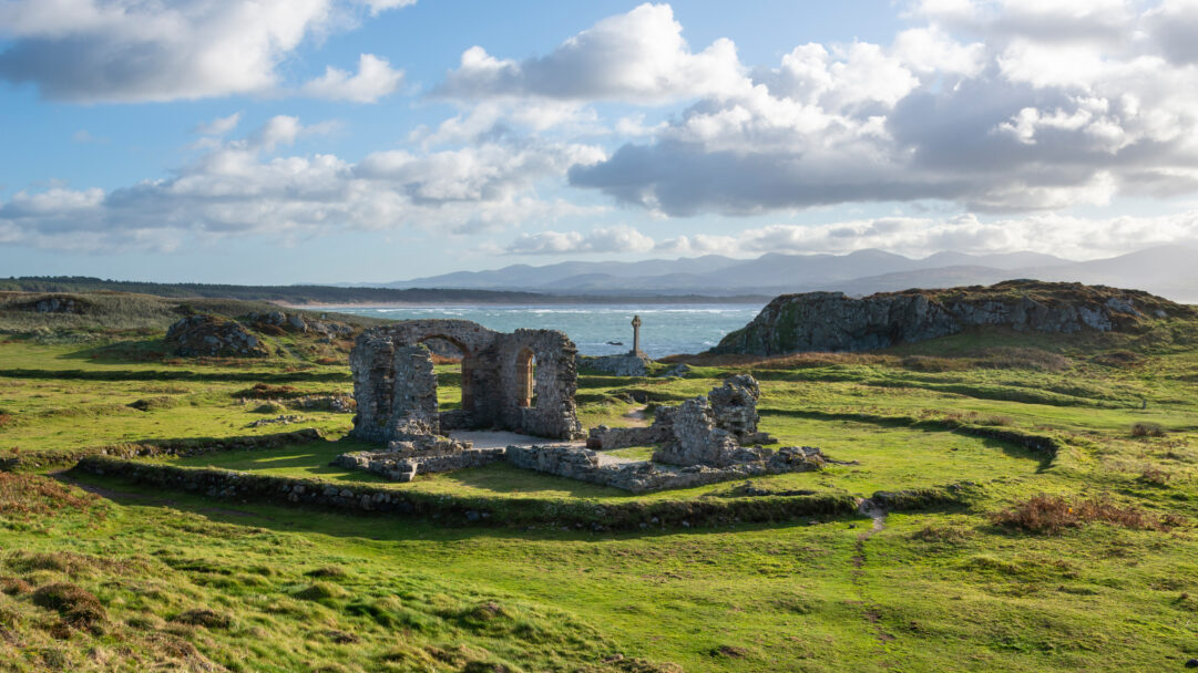 An evocative 4K wallpaper presenting the ancient stone ruins of St Dwynwen's Church nestled in the vibrant green coastal landscape of Ynys Llanddwyn, Wales. Brilliant sunlight dramatically highlights the historic stones and verdant fields, creating a sense of serene grandeur against the windswept sea and distant, hazy mountains.