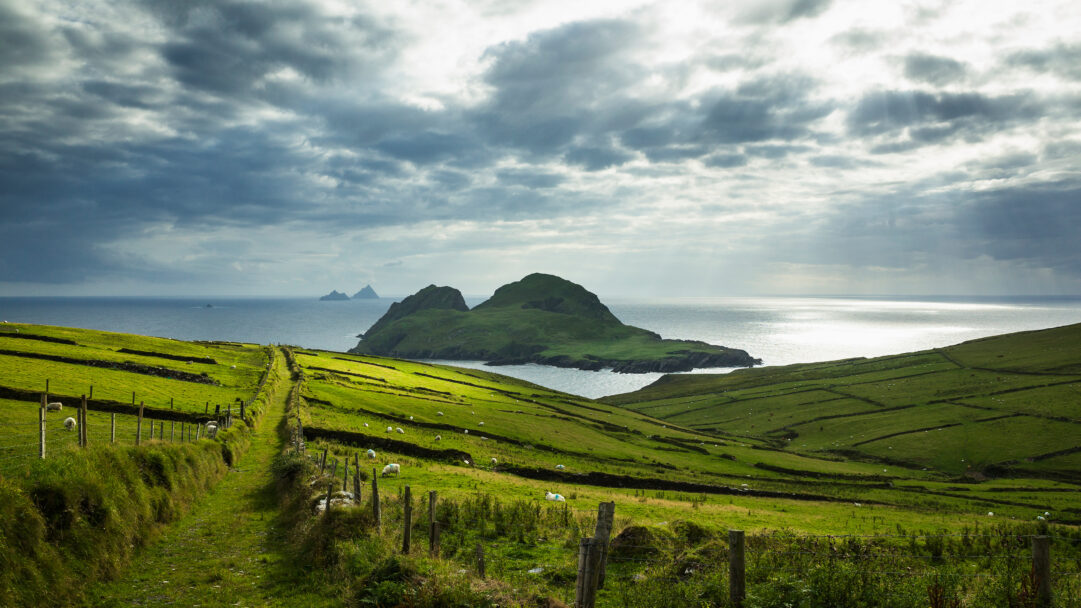 A breathtaking 4K wallpaper depicting the majestic St. Finian's Bay seascape in County Kerry, Ireland, with the iconic Skellig Islands prominent on the horizon, viewed over lush green rolling hills. Dramatic, cloud-strewn skies break open to allow brilliant sunbeams to illuminate the sparkling ocean and distant islands, creating a serene and powerful mood.