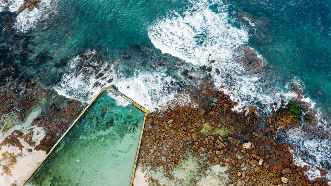 A captivating 4K wallpaper showcasing the St. James Tidal Pool in Cape Town, South Africa, from an aerial ocean view. The vibrant turquoise water of the pool, where a few people swim, sharply contrasts with the wild, white-foamed ocean crashing against the rugged, rust-toned coastline.