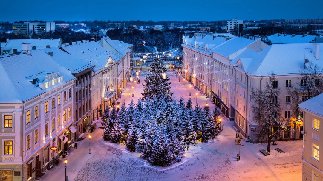 An enchanting 4K wallpaper captures Tartu Town Hall Square, completely blanketed in snow on a serene winter evening. The square comes alive with the warm glow of Christmas lights on the central tree and strung across buildings, creating a festive contrast with the cool blue twilight and snow-covered ground.