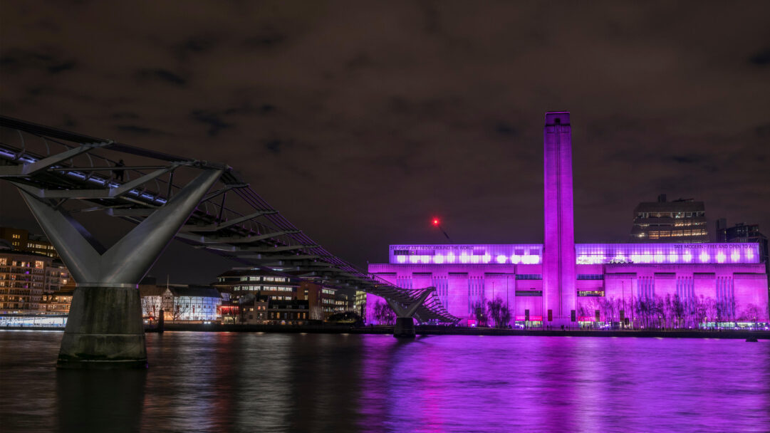 A resplendent 4K wallpaper illuminates the Tate Modern in London, brilliantly lit with purple lights at night, with the Millennium Bridge arcing into the foreground over the River Thames. This vibrant purple glow, celebrating International Women's Day, casts dramatic reflections across the dark water, creating a powerful and commemorative atmosphere.