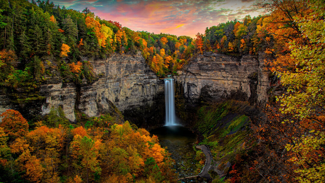 A glorious 4K wallpaper featuring Taughannock Falls State Park in New York, where a powerful waterfall plunges into a deep gorge surrounded by vibrant autumn foliage. The long exposure of the waterfall creates a silky white column, beautifully contrasted by the fiery red and orange hues of the surrounding trees and the dramatic, colorful sky above.