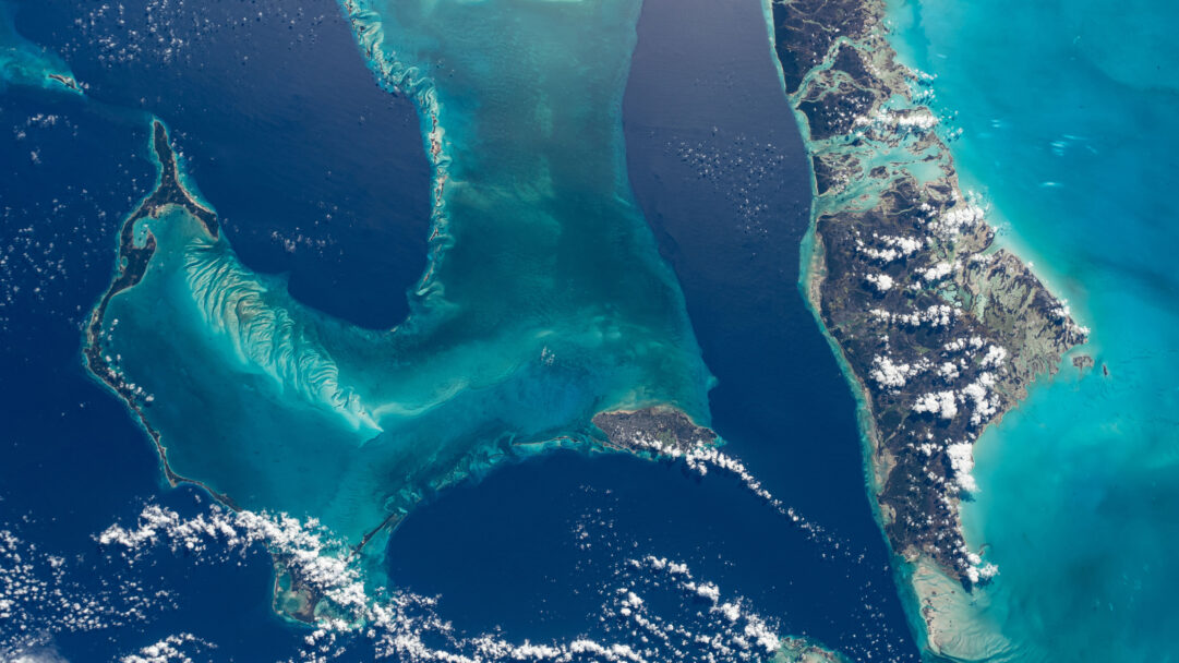 A breathtaking 4K wallpaper revealing the intricate patterns of the Bahamas archipelago and surrounding Atlantic Ocean, as viewed from the International Space Station. The scene highlights a dramatic interplay of deep indigo ocean and luminous turquoise shallow banks, revealing stunning geological contours under scattered white clouds.
