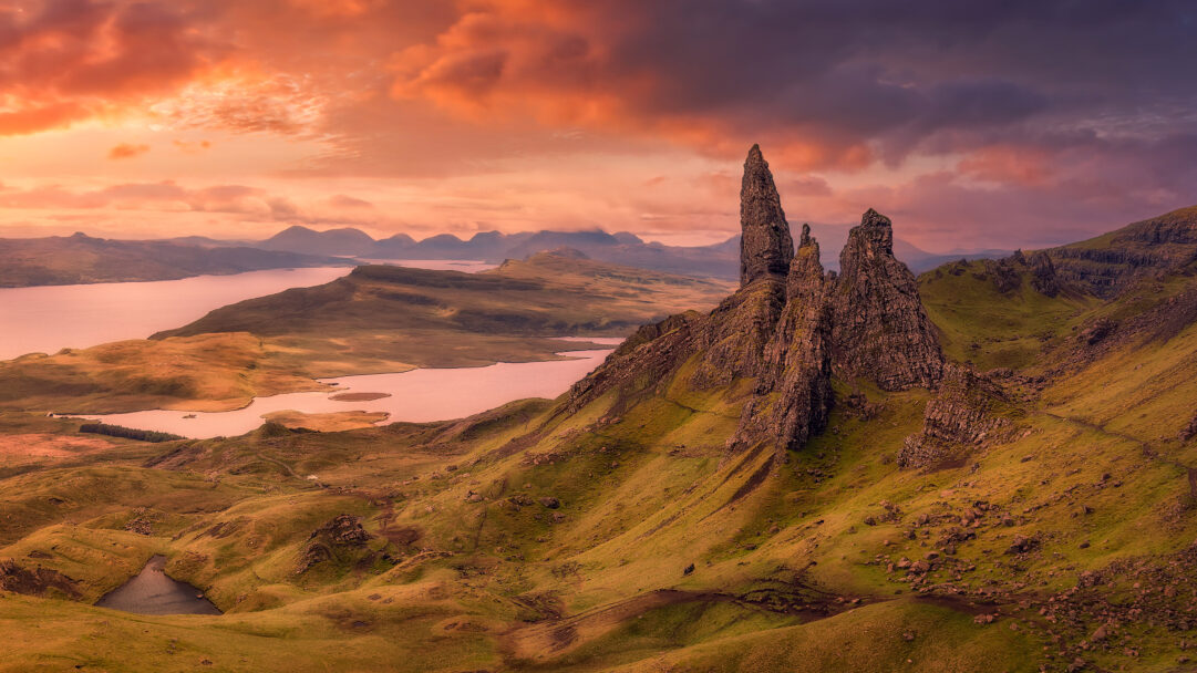 A dramatic 4K wallpaper showcasing The Storr Rock Outcrop, a cluster of rugged peaks, standing proudly on a green hillside overlooking a tranquil loch on the Isle of Skye, Scotland. The scene is bathed in the warm, ethereal glow of a sunset, where vibrant orange and deep purple clouds paint the sky, casting a serene yet powerful atmosphere over the ancient landscape.