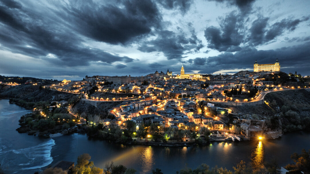 A captivating 4K wallpaper of the historic Toledo, Spain, showcasing its illuminated night cityscape curving majestically around the Tagus River. Golden city lights sparkle against the deep blue water of the river, while a dramatic, cloud-filled twilight sky adds an evocative mood to the ancient Spanish city.