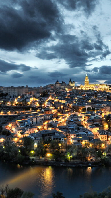 A captivating 4K wallpaper of the historic Toledo, Spain, showcasing its illuminated night cityscape curving majestically around the Tagus River. Golden city lights sparkle against the deep blue water of the river, while a dramatic, cloud-filled twilight sky adds an evocative mood to the ancient Spanish city.